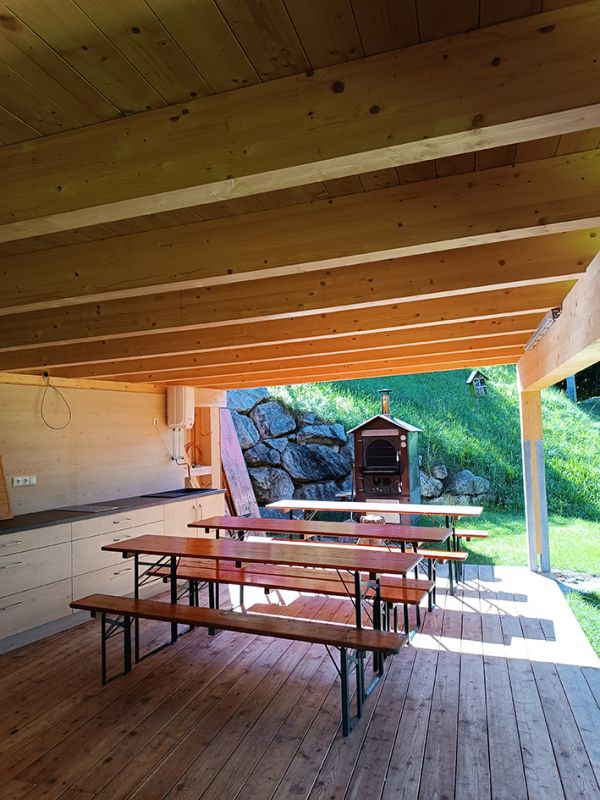 In einem halboffenen Holzpavillon mit Blick ins Freie stehen mehrere Garinturen Biertische und Bänke, auf der linken Seite befindet sich eine Küchenzeile. Direkt am Pavillon steht ein Pizzaofen.