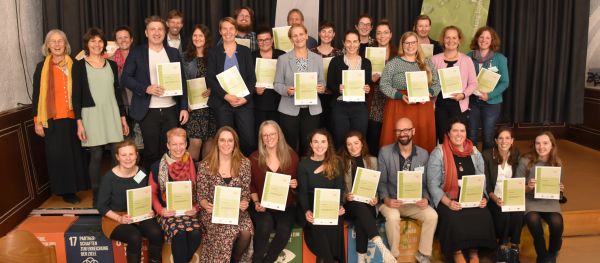Gruppenfoto mit rund 30 Absolvent:innen der Ausbildung mit Zeugnis. 1. Reihe sitzend, die 2 Reihe stehend dahinter