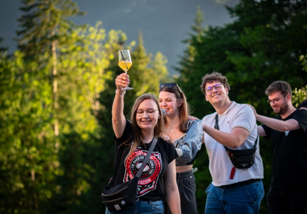 vier junge Menschen tanzen sommerlich gekleidet in einer Polonaise, die junge Frau ganz vorne hält freudig ein gefülltes Sektglas hoch