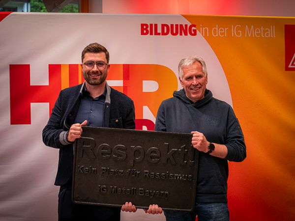 Vor einem Banner der IG Metall halten zwei Männer ein Metallschild mit der Aufschrift "Respekt" in die Kamera