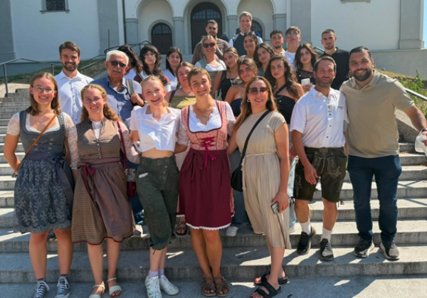 Die rund 30 Teilnehmenden am internationalen Jugendaustausch, einige davon in bayerischer Tracht, haben sich zum Guppenfoto auf einer Treppe im aufgestellt und lächeln in die Kamera