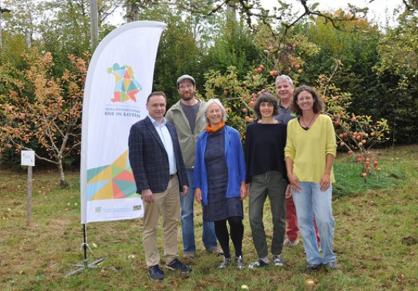 Drei Frauen und drei Männer stehen zum Gruppenfoto auf einer Wiese vor Apfelbäumen neben der ANU-beachflag