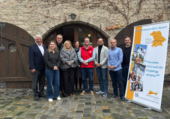 Gruppenfoto von Vertreter:innen aus Politik und Jugendarbeit, im Hintergrund die Burgmauern von Burg Hoheneck, seitlich rechts das Roll-up der Jugendbildungsstätten Bayern