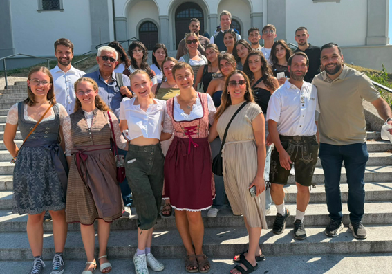 Die rund 30 Teilnehmenden am internationalen Jugendaustausch, einige davon in bayerischer Tracht, haben sich zum Guppenfoto auf einer Treppe im aufgestellt und lächeln in die Kamera