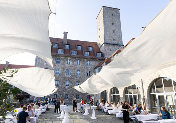 Einige Menschen auf einem festlich geschmückter Platz mit Stehtischen und Tischen und Bänken mit weißen Hussen. Dahinter eine Burg mit einem viereckigen Turm.