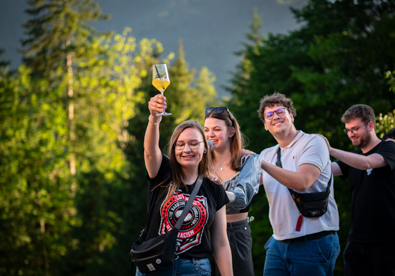 vier junge Menschen tanzen sommerlich gekleidet in einer Polonaise, die junge Frau ganz vorne hält freudig ein gefülltes Sektglas hoch